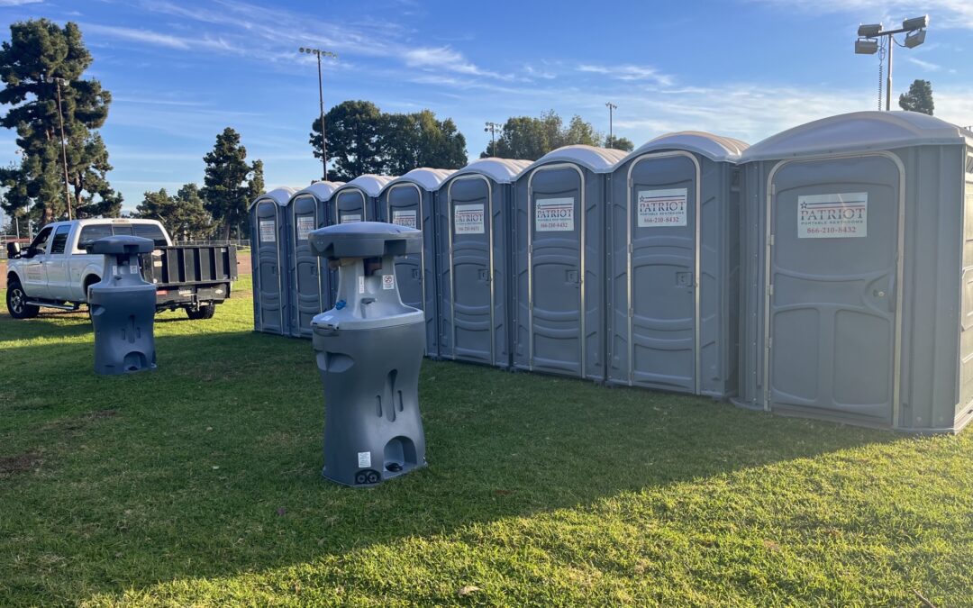 Row of gray portable toilets and handwashing stations on grass beside a parked truck.