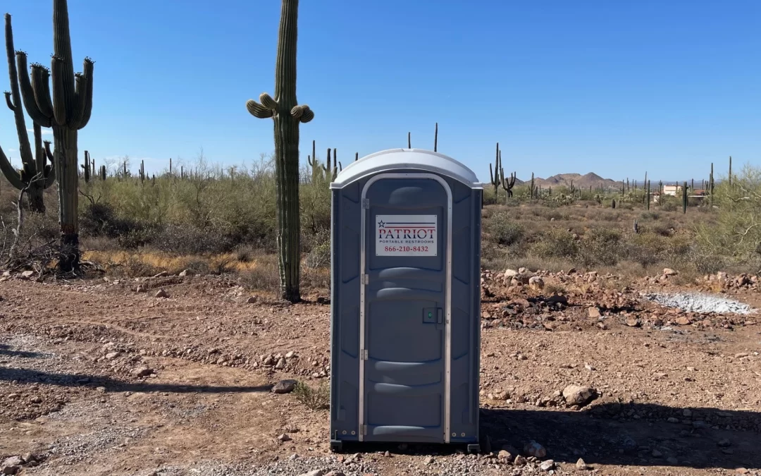Portable restroom placed at a desert construction site with cacti and mountains under clear blue skies.
