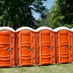 Row of bright orange portable toilets lined up on grass with tall green trees in the background.