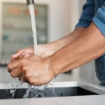 Close-up of hands being washed under a faucet with water splashing into a modern indoor sink.