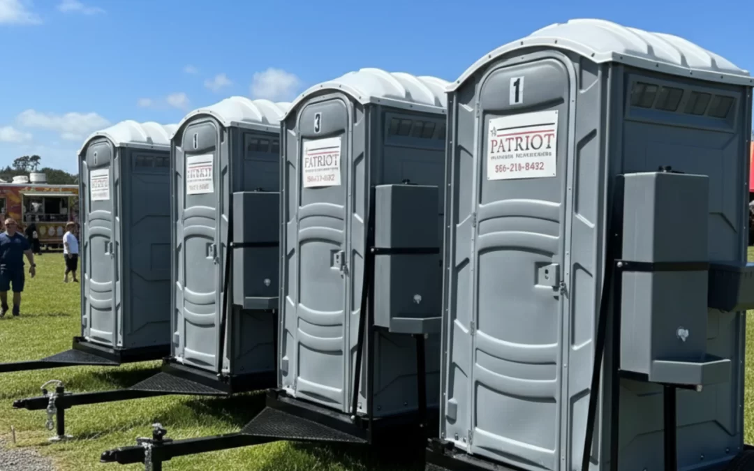 Row of gray portable toilets lined up at an outdoor event with food trucks and people nearby