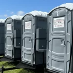 Row of gray portable toilets lined up at an outdoor event with food trucks and people nearby
