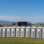 Festival crowd gathers beyond a line of gray portable toilets facing a distant concert stage