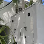 White portable restroom trailer beside tropical plants and a modern home in bright daylight outside.