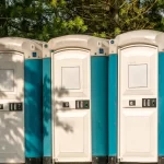 Three teal and white portable toilets tucked among pine trees in warm sunlight outdoors